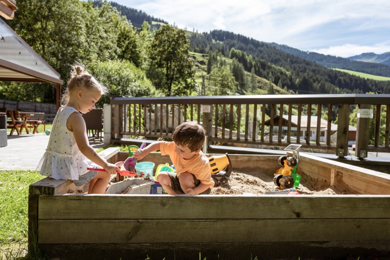 Zwei Kinder spielen in einem Sandkasten vor einer malerischen Berglandschaft