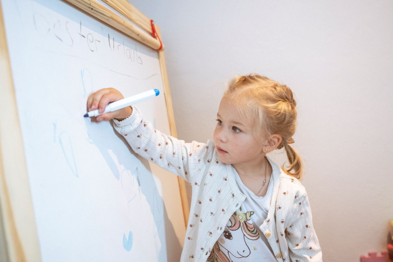 Ein kleines M&auml;dchen schreibt mit einem Stift auf eine wei&szlig;e Tafel im Kinderspielzimmer im Familienhotel Bachschmied.