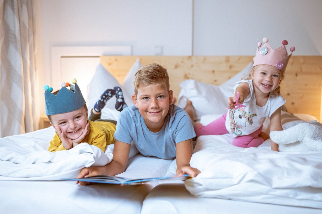 Drei lachende Kinder mit Kronen liegen auf einem Bett und schauen ein Buch an im Hotel Bachschmied.
