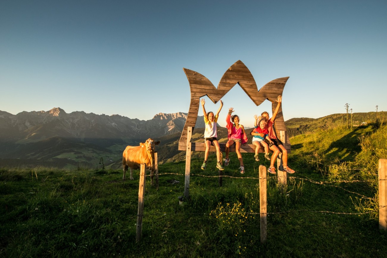 Vier Kinder sitzen auf einem Holzrahmen der Hochk&ouml;nigskrone vor der Bergkulisse, daneben steht eine Kuh. &copy;TVB_Hochk&ouml;nig-Schartner