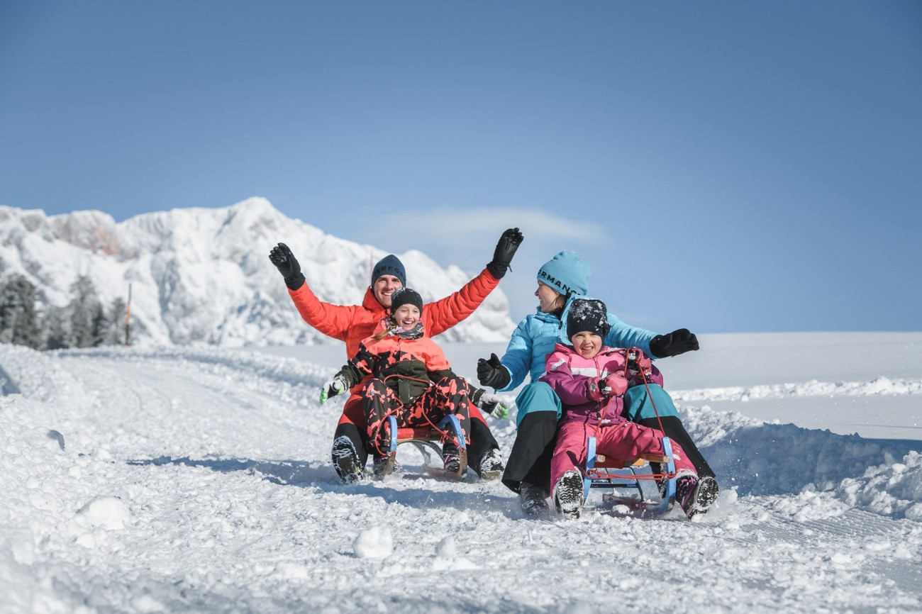 Eine Familie rodelnd auf einer schneebedeckten Piste vor malerischen Bergen im Skiurlaub am Hochk&ouml;nig. &copy;TVB_Hochk&ouml;nig