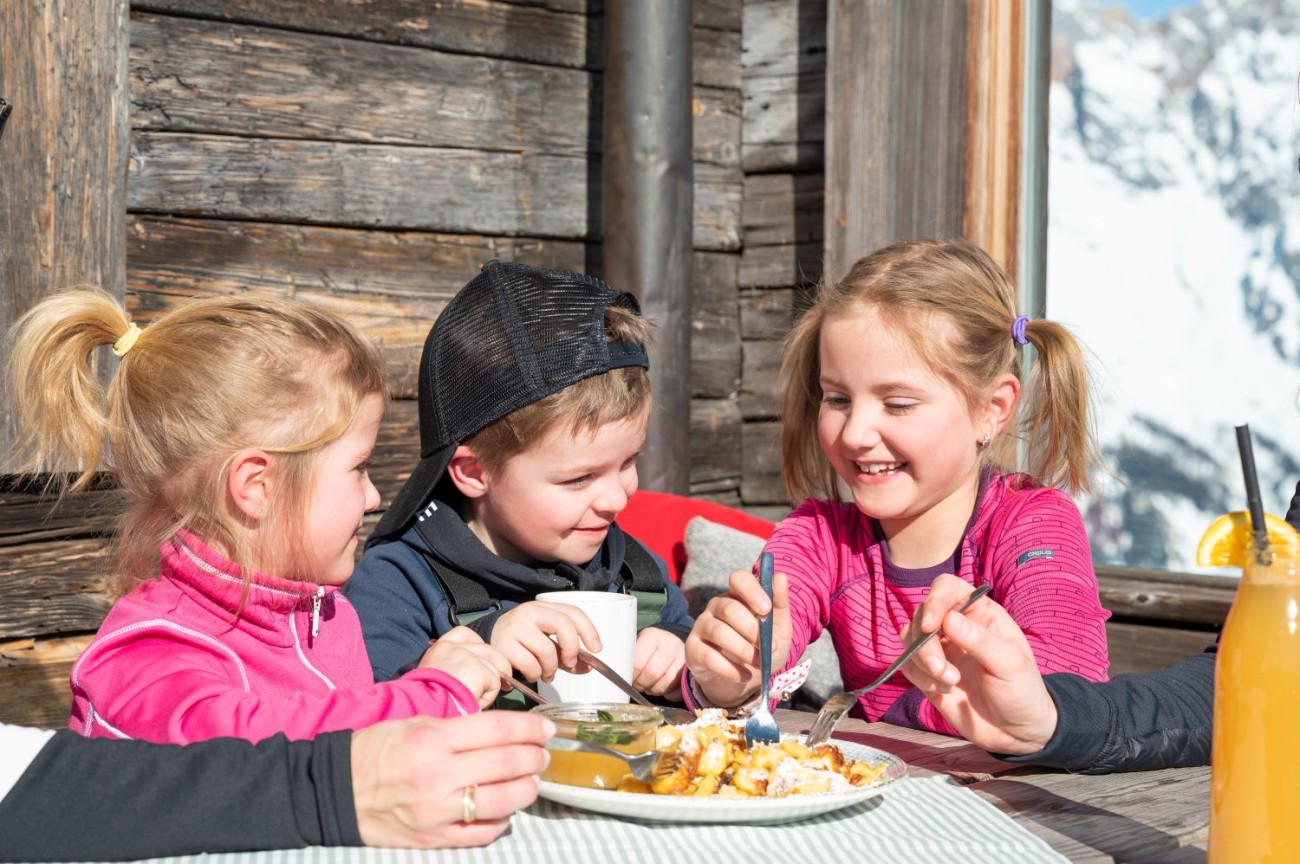 Drei lachende Kinder sitzen drau&szlig;en und essen gemeinsam an einem Holztisch im Familienurlaub im Hotel Bachschmied. &copy;TVB Hochk&ouml;nig