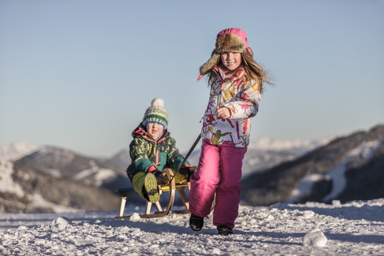 Zwei Kinder rodeln fr&ouml;hlich im Schnee vor einer Berglandschaft im Familienurlaub in Maria Alm. &copy;TVB Hochk&ouml;nig