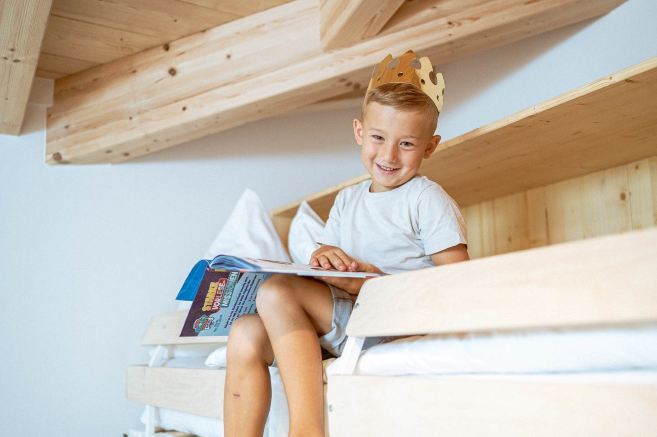 Ein l&auml;chelnder Junge mit Krone liest ein Buch auf einem Hochbett im Familienurlaub im Hotel Bachschmied.