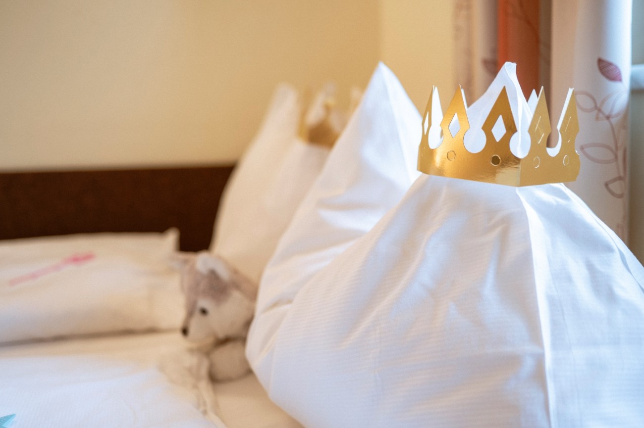 Wei&szlig;e Kissen mit goldenen Papierkronen und ein Pl&uuml;schtier auf einem Bett.